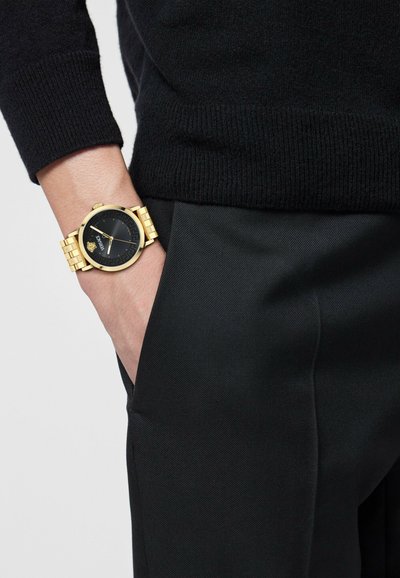 Kuldne käekell musta kellaga, millel on tekstureeritud pind ja kuldne link-randmestik, kantud tumeda sviidri ja mustade pükstega.