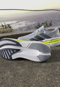 Zapatillas deportivas de color gris claro con rayas grises, un acento amarillo, material superior texturizado y una suela con un patrón en blanco y negro.