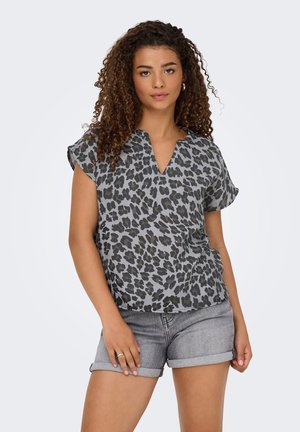 Femme aux cheveux bouclés portant un chemisier gris et noir à motif léopard et un short en jean gris retroussé, debout devant un fond uni.
