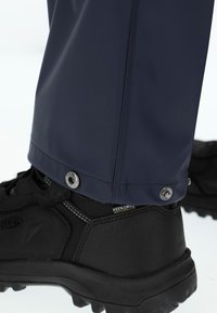 Czarny wodoodporny but turystyczny częściowo zakryty granatowymi spodniami z zatrzaskami na dole.