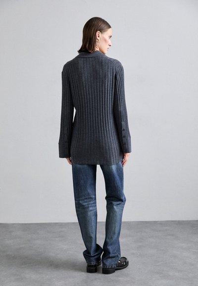 Cardigã cinza canelado com mangas longas, apresentando detalhes em botão nos punhos, combinado com jeans azul. Textura suave, corte ajustado.
