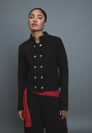 Femme debout sur un fond gris, portant une veste noire avec des boutons argentés, un pantalon large noir, un pull rouge noué à la taille et des chaussures blanches.