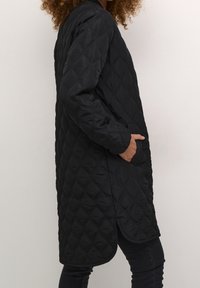 Person mit lockigem Haar, der eine schwarze, gesteppte lange Jacke und schwarze Hosen trägt, mit einer Hand in der Tasche, steht vor einer schlichten hellen Wand.