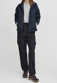 Persona che indossa una giacca blu marina sopra una camicia grigio chiaro, pantaloni cargo neri e scarpe da trekking marroni, in piedi con le mani nelle tasche.