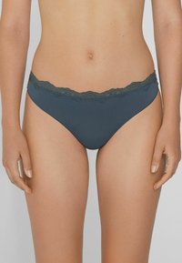 Femme portant un sous-vêtement de style bikini gris foncé avec des bordures en dentelle, se tenant debout avec les mains détendues le long du corps sur un fond uni.