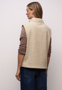 Beżowa kamizelka z polaru z wysokim kołnierzem, o fakturowanej powierzchni, bez rękawów. Noszona na brązowej koszuli z długim rękawem i połączona z niebieskimi dżinsami.