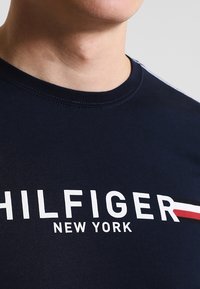 Námořnická modrá bavlněná tričko s kulatým výstřihem, na kterém je bílé logo "TOMMY HILFIGER NEW YORK" a červený akcentový pruh na rameně.