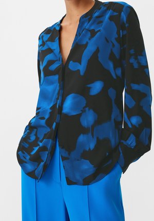 Kvinde iført en sort bluse med lyseblå abstrakte blomsterprint og matchende lyseblå bukser med hænderne i lommerne.