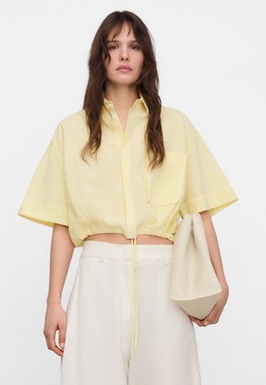 Femme aux longs cheveux bruns porte une chemise courte jaune rayée boutonnée, un pantalon blanc taille haute et tient un sac fourre-tout beige.