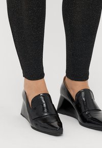 Juodos patentinės wedges su smailiu nosiuku ir lygiu, blizgiu paviršiumi. Derinamos su juodomis tamprėmis, kuriose yra sidabrinių blizgučių.