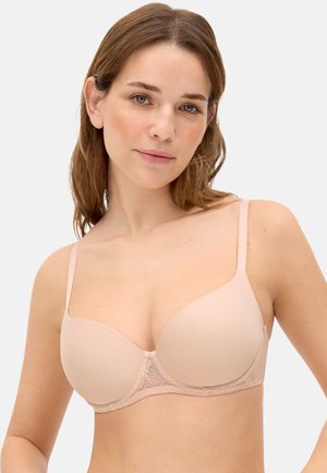 Mujer de piel clara y cabello castaño vistiendo un sujetador color nude, ligeramente acolchado y con detalles de encaje, sobre un fondo blanco liso.