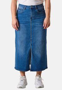 Denim maxi skirt with a front slit, blue wash, high waist, five pockets, and button closure. Paired with white athletic shoes.