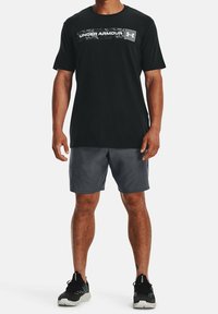 T-shirt nera a maniche corte con logo "Under Armour" riflettente, abbinata a pantaloni corti grigio scuro. L'uomo indossa scarpe da ginnastica nere.