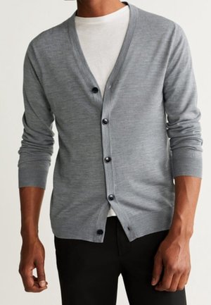 Uomo con un cardigan grigio chiaro abbottonato sopra una maglietta bianca e pantaloni neri, in piedi con le braccia rilassate.