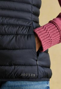 Melnā puffa veste ar horizontālu šuvju dizainu, kurai ir rāvējslēdzēja kabata un 'JOULES' logs. Rozā adītas piedurknes ar ribotu tekstūru.