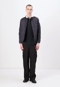 Veste noire matelassée avec un col montant, dotée d'une fermeture éclair à l'avant et de poches latérales, portée sur un polo noir et un pantalon cargo.