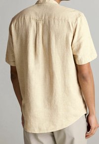 Chemise à manches courtes en tissu texturé beige clair, avec un pli au dos, un ourlet droit et une coupe décontractée. Agrémentée de tons doux.