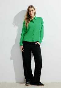 Camicia verde a maniche lunghe con bottoni davanti, abbinata a pantaloni neri larghi e scarpe scure. Il tessuto appare leggero e morbido.