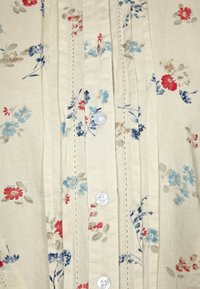 Tissu crème avec de petits motifs floraux rouges, bleus et beiges, présentant des plis pintuck verticaux et des boutons blancs au centre.