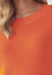 Persona bionda che indossa un maglione arancione a costine con scollo rotondo, che mostra la parte superiore del petto e delle spalle.