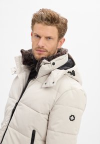 Lichtgewicht pufferjack in crème met een bontkraag, ritssluiting aan de voorkant en twee zijzakken. Zwarte accenten en logo op de linkermouw.