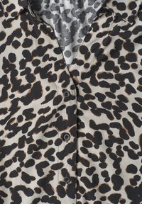 Gros plan d’une chemise beige avec un motif léopard irrégulier noir et marron et des boutons noirs sur la patte de boutonnage avant.
