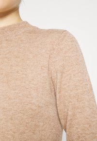 Pull en tricot marron clair avec un col rond et une apparence texturée. Le tissu présente une subtile variation de couleur et de détails de points.