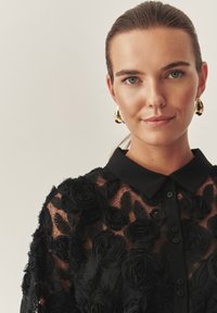 Blusa nera con texture in pizzo floreale, dotata di colletto, chiusura a bottoni e applicazioni di rose 3D, completata da orecchini a cerchio dorati.