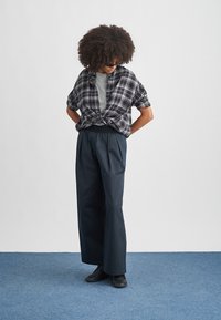 Svartvit rutig skjorta med kort ärm knuten i midjan över en grå rundhalsad tröja, kombinerad med marinblå vida byxor och svarta skor.