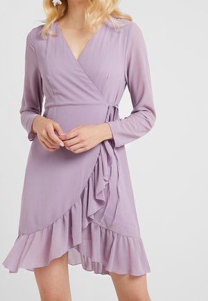 Femme portant une robe portefeuille à manches longues, couleur violet clair, avec des détails à volants et une ceinture nouée sur le côté à la taille, debout devant un fond uni.