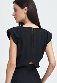 Czarna bluzka z krótkimi rękawami z naszywką, z zapięciem na zamek z tyłu i małym wycięciem. Gładki materiał z dopasowanym krojem.