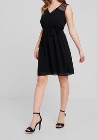 Nainen, jolla on yllään musta hihaton mekko, jossa on pitsiset olkapääkoristukset ja sidottu vyö, yhdistettynä mustiin korkokenkiin.