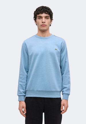 Jonge man staat, draagt een lichtblauwe trui met ronde hals en een klein groen logo op de borst en zwarte broek.