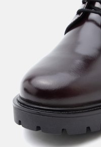 Γυαλιστερό μπορντό δερμάτινο παπούτσι με στρογγυλή μύτη, μαύρη υφασμάτινη σόλα και αντίθετη ραφή κατά μήκος της άκρης.