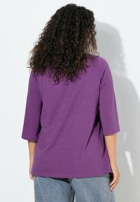 Top morado de manga tres cuartos con un ajuste holgado, confeccionado en una tela suave y texturizada; la parte trasera es lisa con un dobladillo ligeramente curvado.