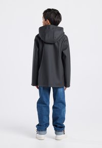 Black waterproof jacket with a hood, rubber-like texture, oversize fit, paired with light blue denim jeans and white sneakers. Back view.