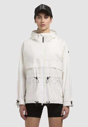 Leichte weiße Jacke mit Kapuze, verstellbaren Kordelzügen an der Taille und den Bündchen, ausgestattet mit zwei seitlichen Reißverschlusstaschen. Schwarze Baseballmütze.