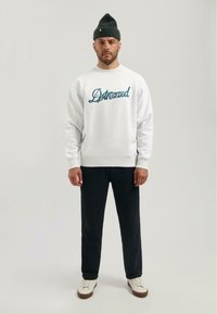 Felpa grigia con scritta blu "Dstressed", abbinata a pantaloni scuri e sneaker bianche. Berretto di lana verde scuro sulla testa.