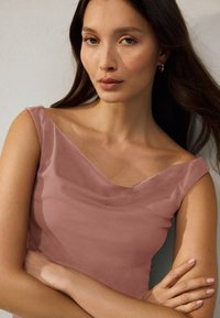 Tânără cu păr lung și închis la culoare, purtând un top roz prăfuit fără mâneci, cu brațele încrucișate și cercei mici de aur în formă de cerc, pe un fundal simplu.