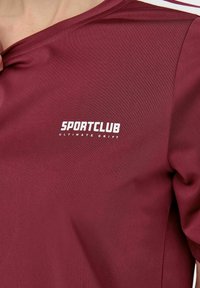 Bordeauxfarbenes Sportshirt mit glatter Textur, mit "SPORTCLUB" und "ULTIMATE DRIFT" in Weiß auf der Vorderseite bedruckt. Einfaches Rundhalsdesign.