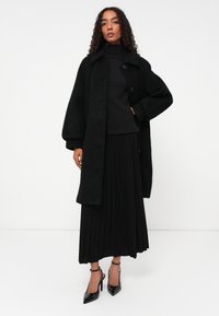 Cappotto in misto lana nero con maniche oversize e grandi bottoni, abbinato a un dolcevita nero aderente e una gonna nera plissettata. Scarpe con tacco alto.