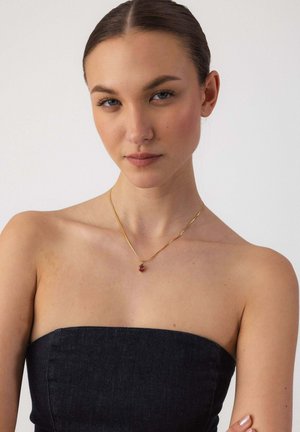 Junge Frau mit zurückgebundenem Haar, die ein trägerloses schwarzes Oberteil und eine goldene Halskette mit einem kleinen roten Herzanhänger trägt.