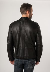 Giacca di pelle nera con una superficie testurizzata, caratterizzata da un colletto alto, cuciture sulle spalle e un design con una fessura sul retro. Abbinata a jeans blu.