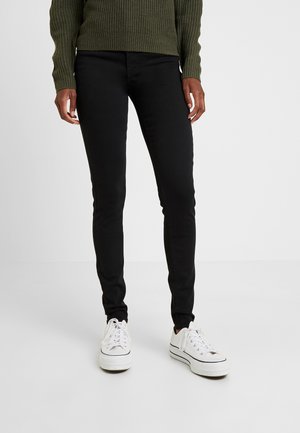 Persoon draagt zwarte slim-fit broek, witte sneakers en een donkergroene gebreide trui, staand op een effen witte vloer.