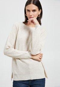 Lichtbeige sweater met een gestructureerde brei, ronde halslijn en subtiele zijsplitten. Lange mouwen met geribde manchetten. Gecombineerd met donkere jeans.
