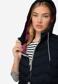 Marinblå pufferväst med zigzag-quilting, grå dragkedja och rosa foder. Har en huva och en randig skjorta under, som hålls öppen av en hand.