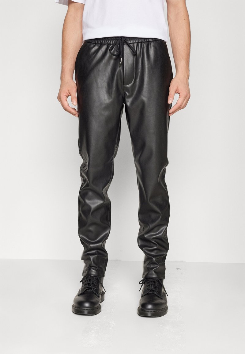 Zwarte faux leren joggers met elastische tailleband, trekkoordsluiting en taps toelopende benen. Gepaard met zwarte veterschoenen.