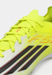 Φωτεινό κίτρινο ποδοσφαιρικό παπούτσι με υφή από συνθετικό υλικό, μαύρες ρίγες και κόκκινες λεπτομέρειες. Εξοπλισμένο με κίτρινα κορδόνια και ελαφρύ σχεδιασμό.