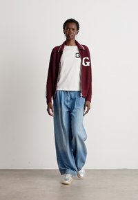 Cardigan a maglia bordeaux con bordi bianchi e logo, indossato sopra una T-shirt bianca. Jeans a gamba larga blu e sneakers grigie completano l'outfit.