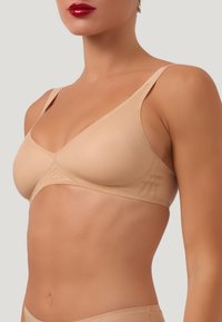 Beige, ribbat tyg-bh med triangulär form, tunna band och en slät yta. Har geometriska design detaljer på bandet.